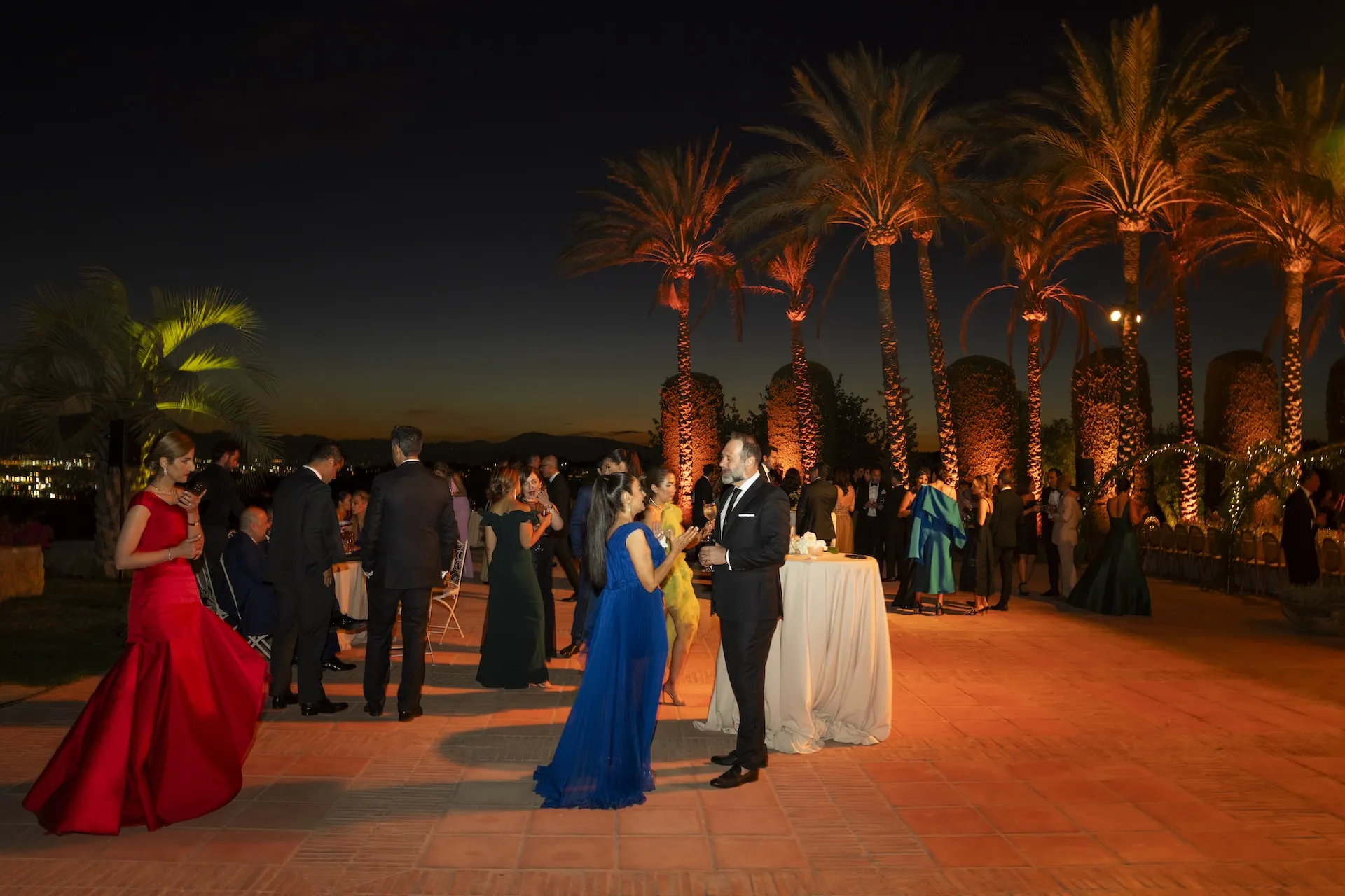 Guests in formal evening attire at an outdoor wedding reception beneath illuminated palm trees